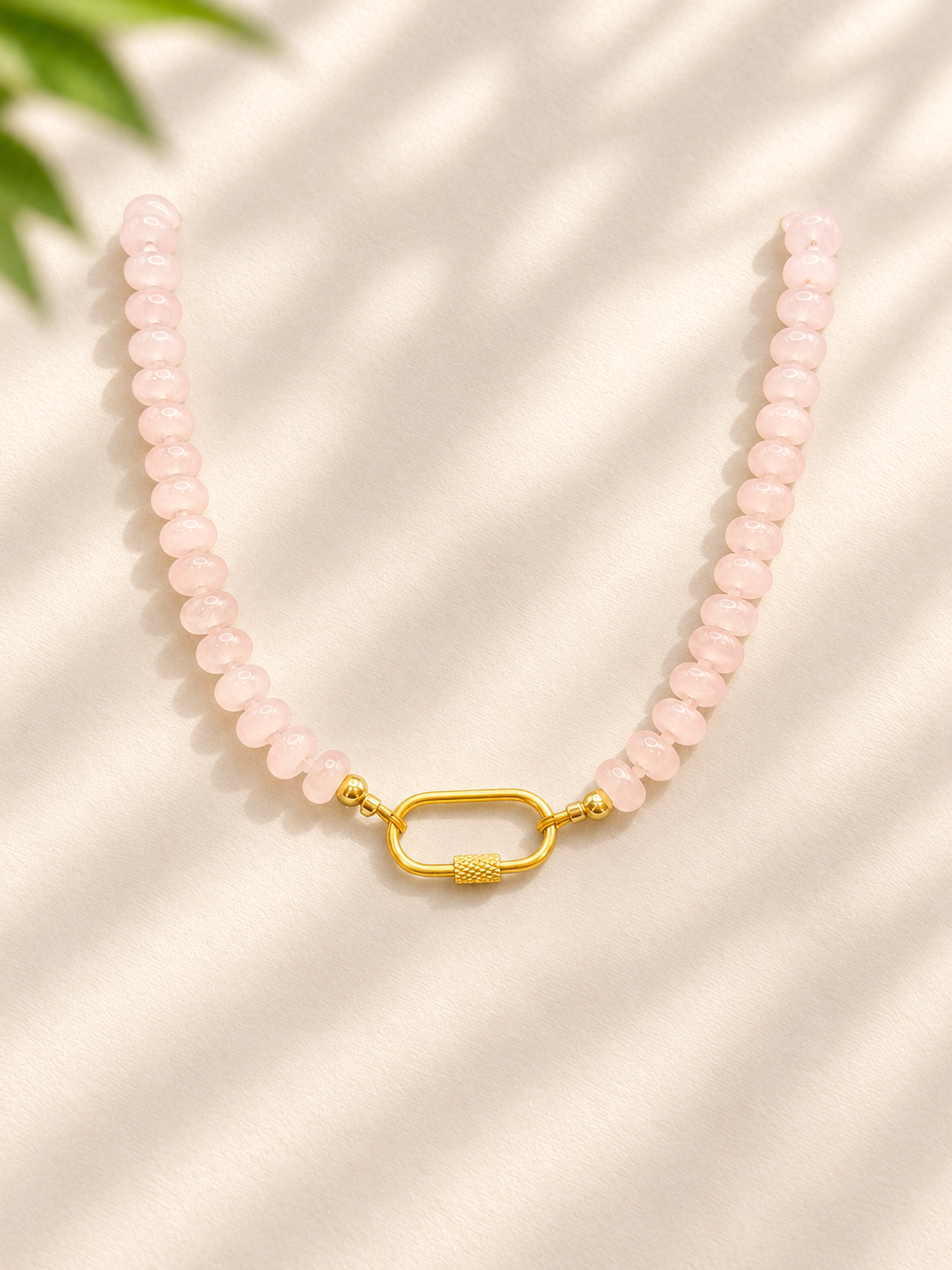 A beaded necklace made of light pink faceted beads with a textured gold clasp, displayed on a cream-colored fabric background with soft leaf shadows.
