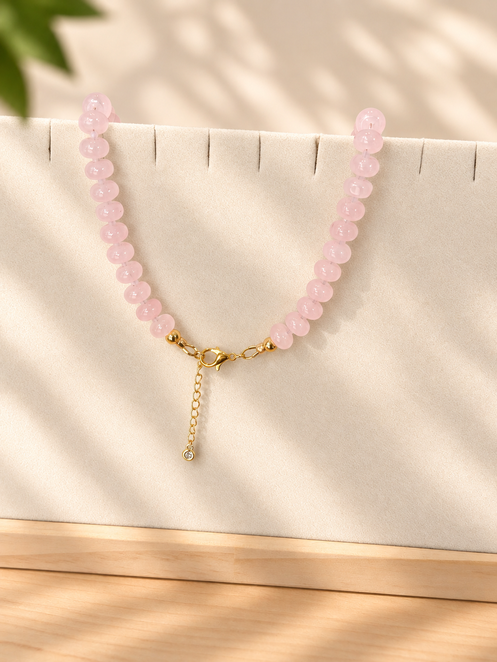 A pink beaded necklace with a gold clasp and a small dangling chain charm, displayed on a cream jewelry stand with soft leaf shadows.