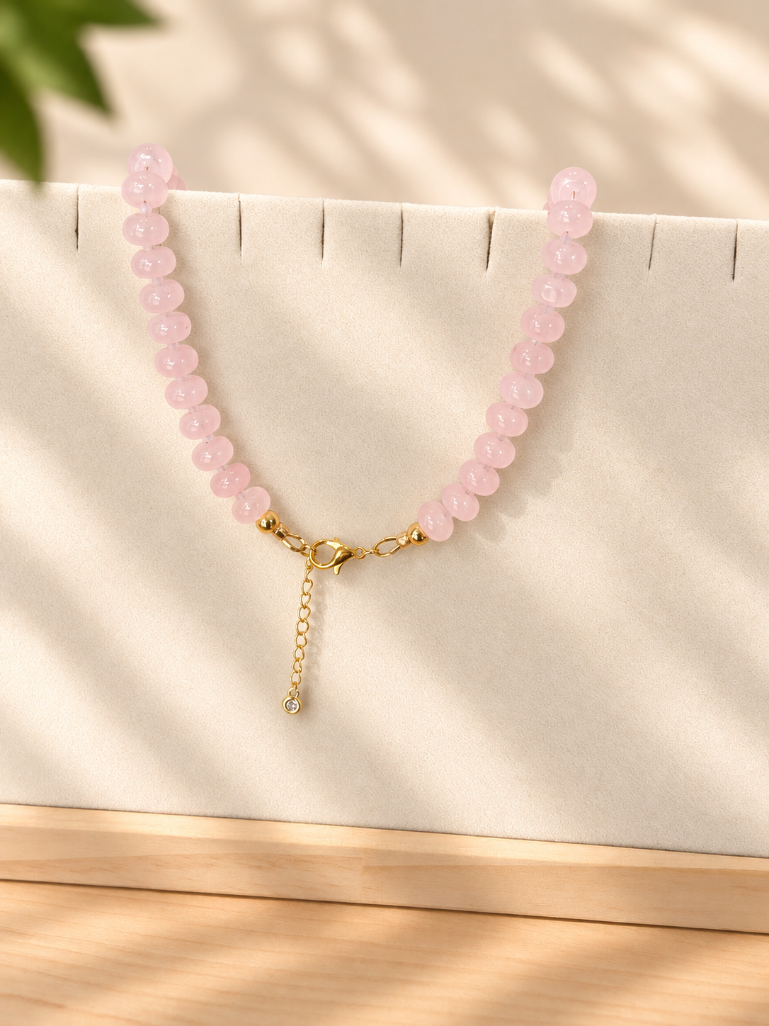 A pink beaded necklace with a gold clasp and a small dangling chain charm, displayed on a cream jewelry stand with soft leaf shadows.