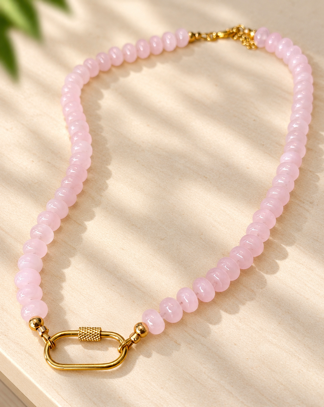A pink beaded necklace with a gold clasp, displayed on a light wood surface with soft leaf shadows.