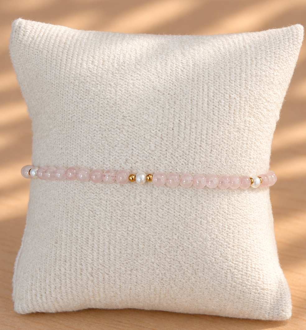 A delicate pink beaded bracelet with small gold and white beads, displayed on a textured cream pillow against a warm wooden background with soft leaf shadows.