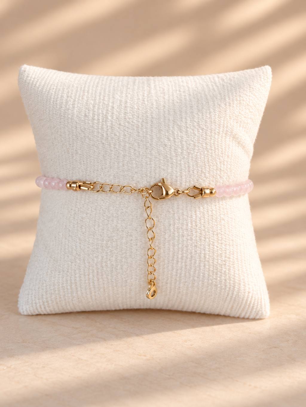 A delicate bracelet with a light pink beaded section and gold-tone chain details, displayed on a textured cream pillow against a warm beige background with soft shadow patterns.