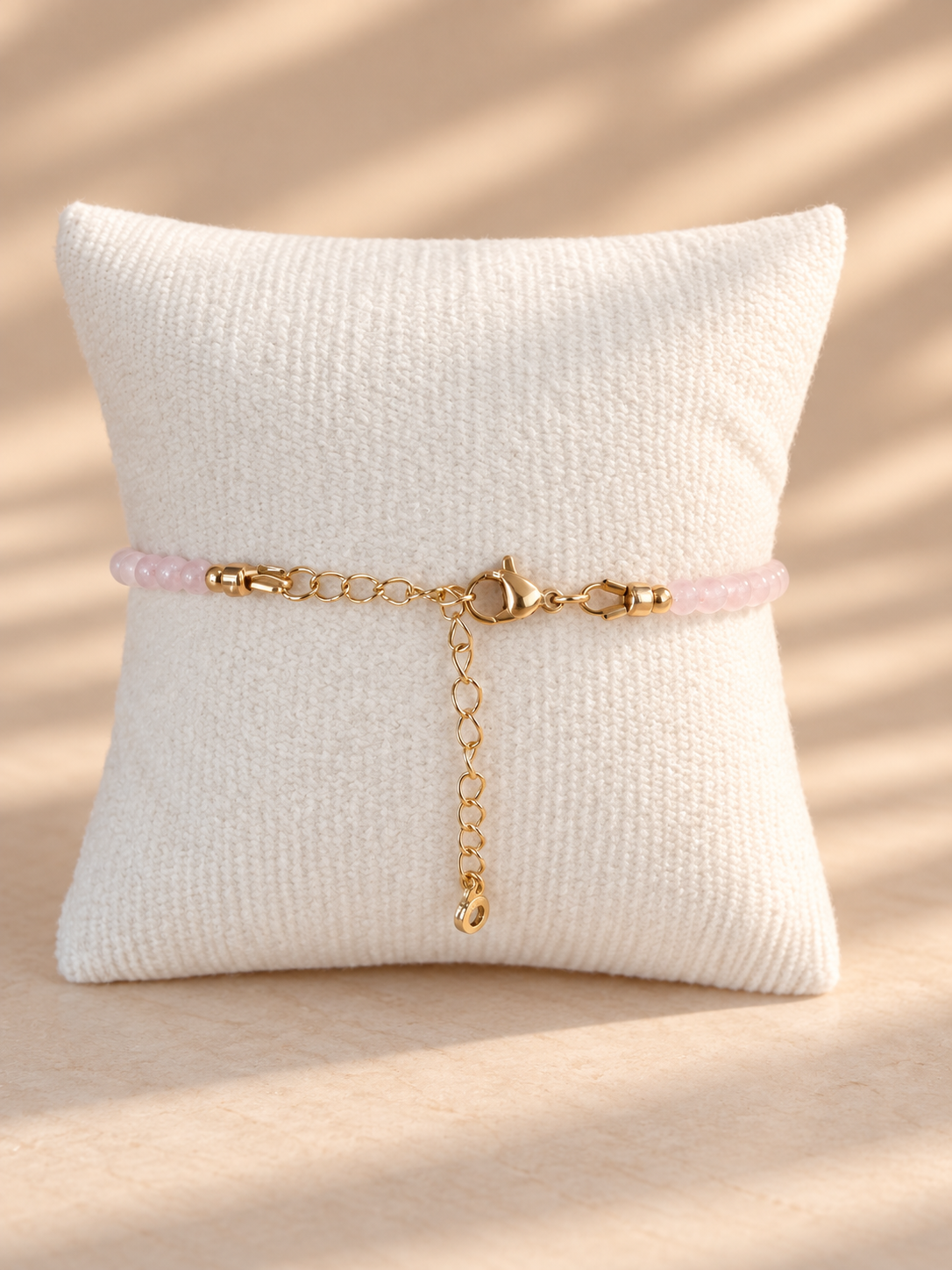 A delicate bracelet with a light pink beaded section and gold-tone chain details, displayed on a textured cream pillow against a warm beige background with soft shadow patterns.