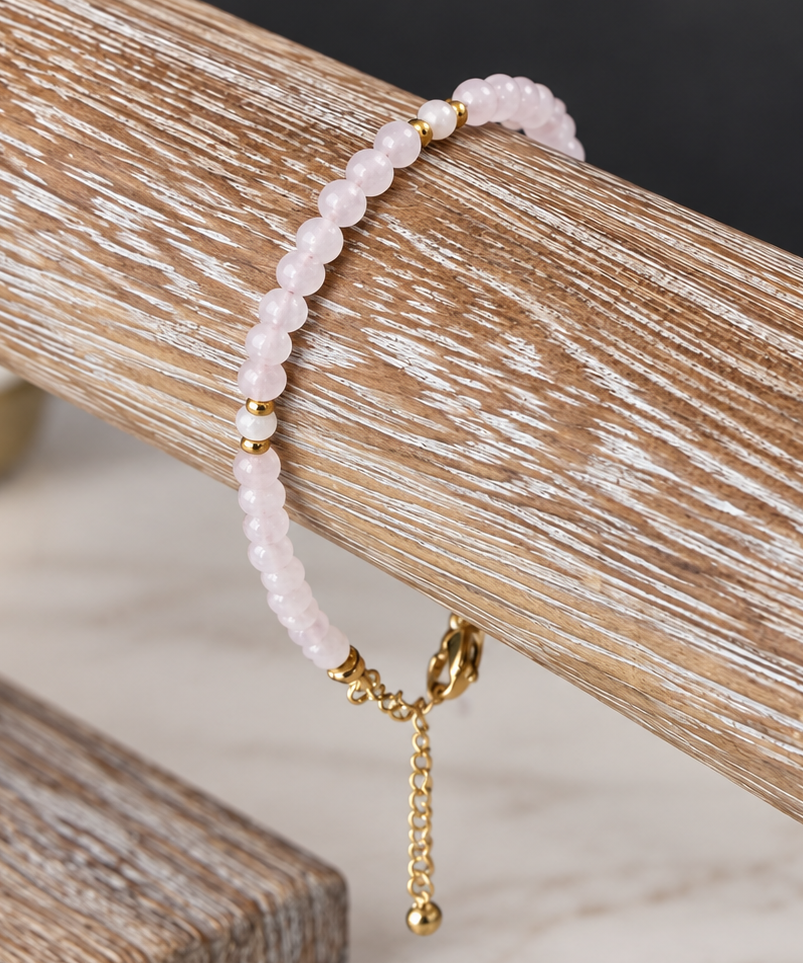 A delicate beaded bracelet in pale pink with small gold spacer beads and a gold clasp, displayed on a rustic wooden jewelry stand.