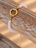 A delicate bracelet with a pale pink beaded section, a gold-tone circular ring clasp, and a small dangling gold chain, displayed on a rustic wooden stand.
