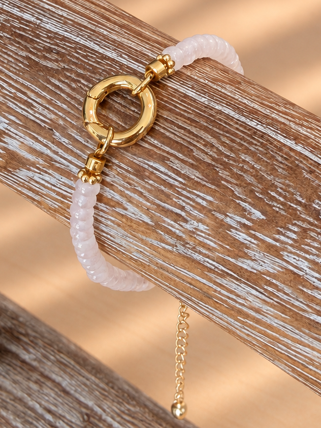 A delicate bracelet with a pale pink beaded section, a gold-tone circular ring clasp, and a small dangling gold chain, displayed on a rustic wooden stand.