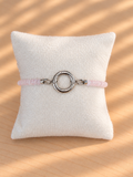 A light pink beaded bracelet with a polished silver-tone circular ring clasp, displayed on a textured white pillow over a light wood surface with soft diagonal shadows.