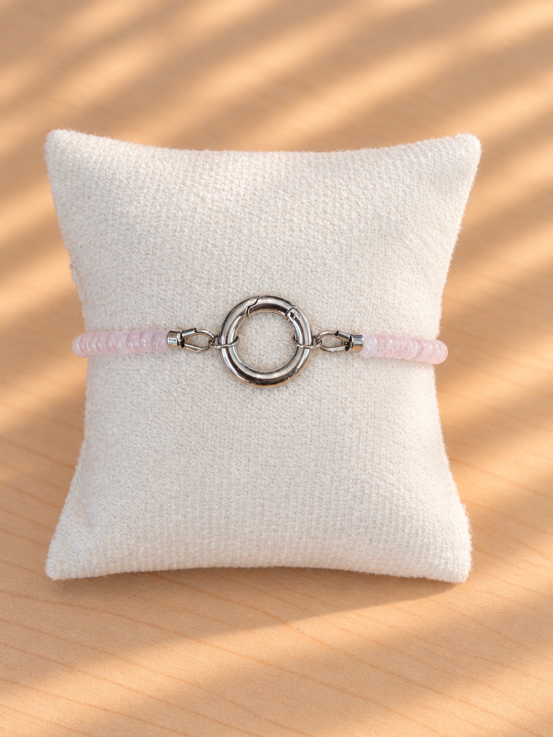 A light pink beaded bracelet with a polished silver-tone circular ring clasp, displayed on a textured white pillow over a light wood surface with soft diagonal shadows.