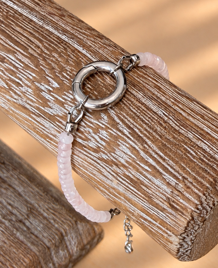 A delicate bracelet with a silver-toned ring clasp, a pale pink beaded section, and a small dangling charm, displayed on a rustic wooden stand against a warm beige background with soft shadow patterns.