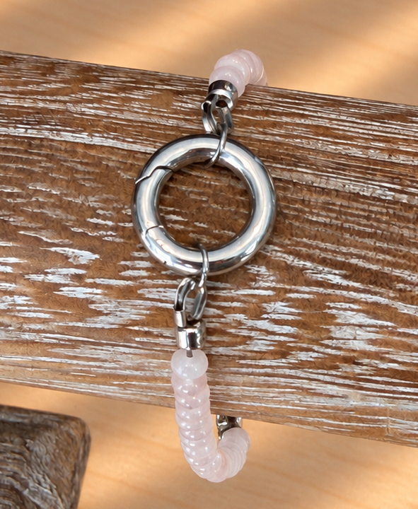A silver-toned ring clasp with a dangling textured pale pink bead charm, displayed on a rustic wooden stand against a sunlit wood surface.