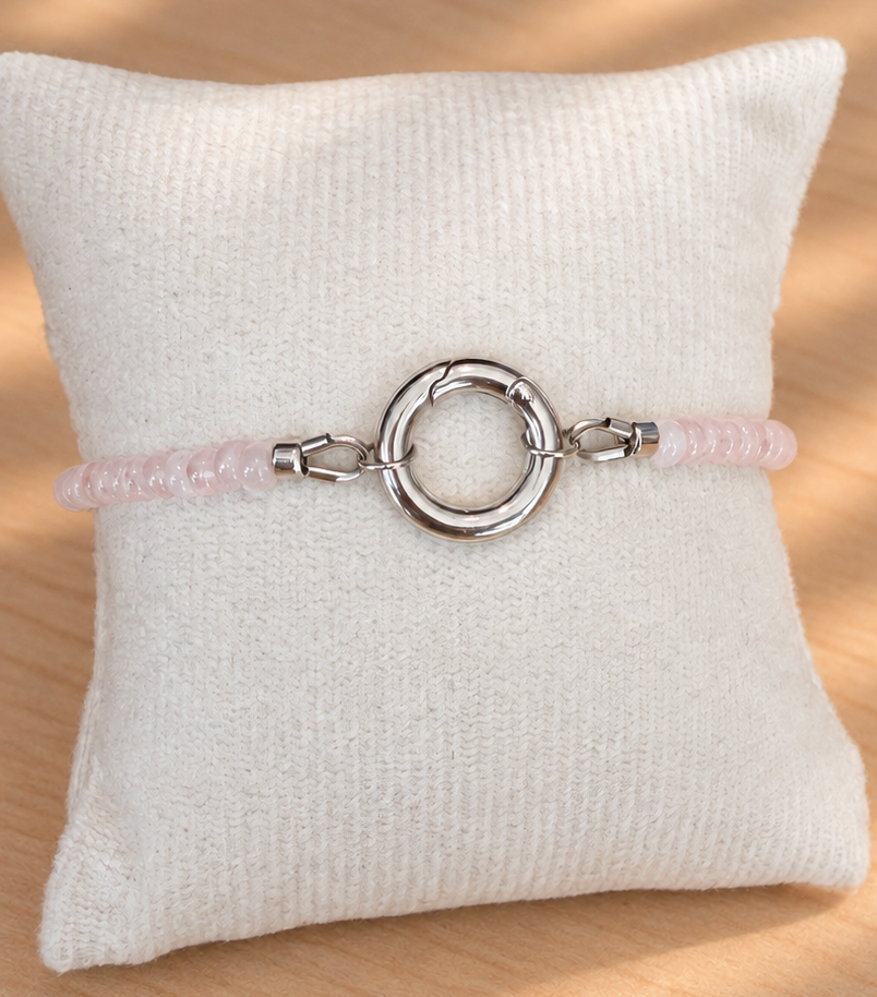 A delicate pink beaded bracelet with a large silver-toned circular ring clasp, displayed on a textured cream fabric pillow over a light wood surface with soft shadow overlays.