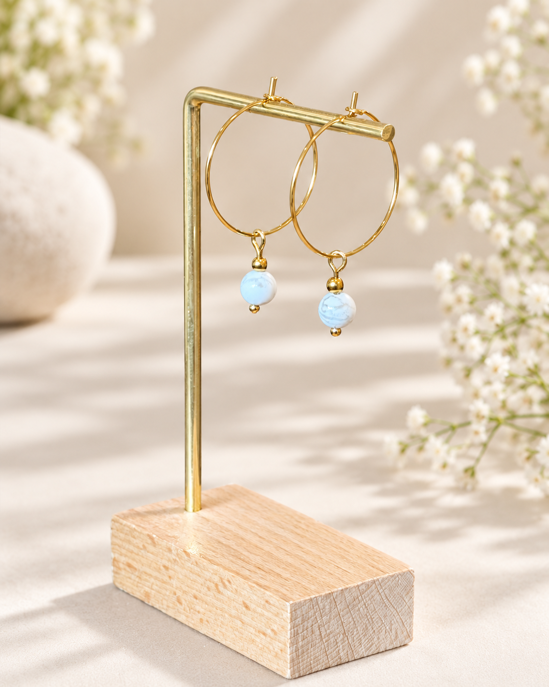 Gold hoop earrings with light blue bead drops displayed on a wooden stand against a softly blurred floral background.