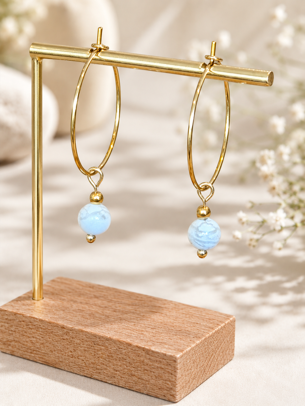 A pair of gold hoop earrings with light blue marbled bead drops, displayed on a gold stand with a wooden base against a soft beige background with blurred white flowers.