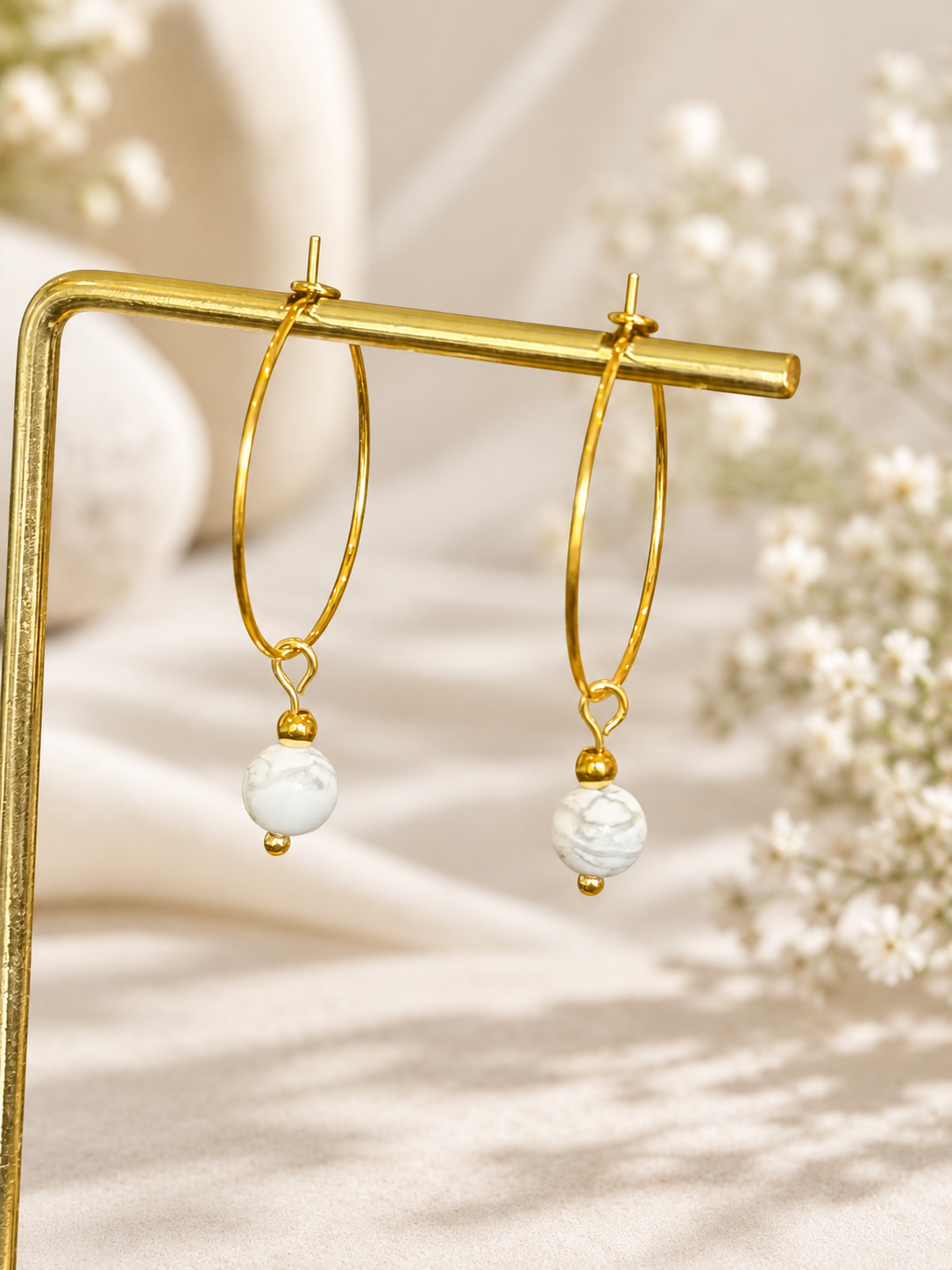 A pair of gold hoop earrings with white marbled bead drops, displayed on a gold stand against a soft beige background with blurred white flowers.