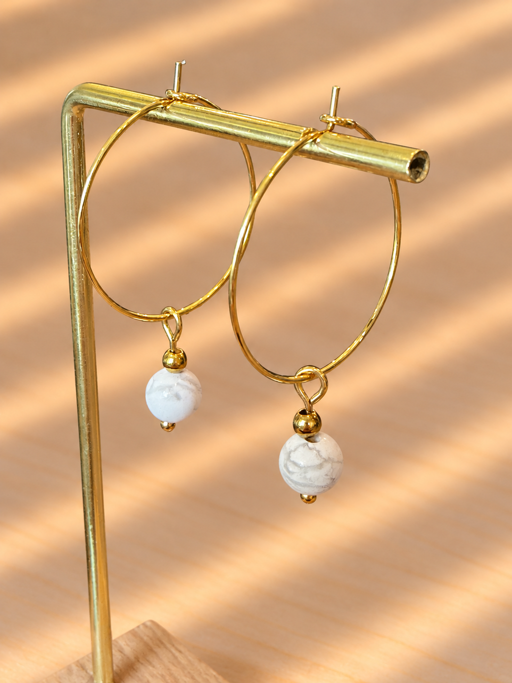 Gold hoop earrings with white marble bead drops, displayed on a gold stand against a warm beige background with soft diagonal shadows.