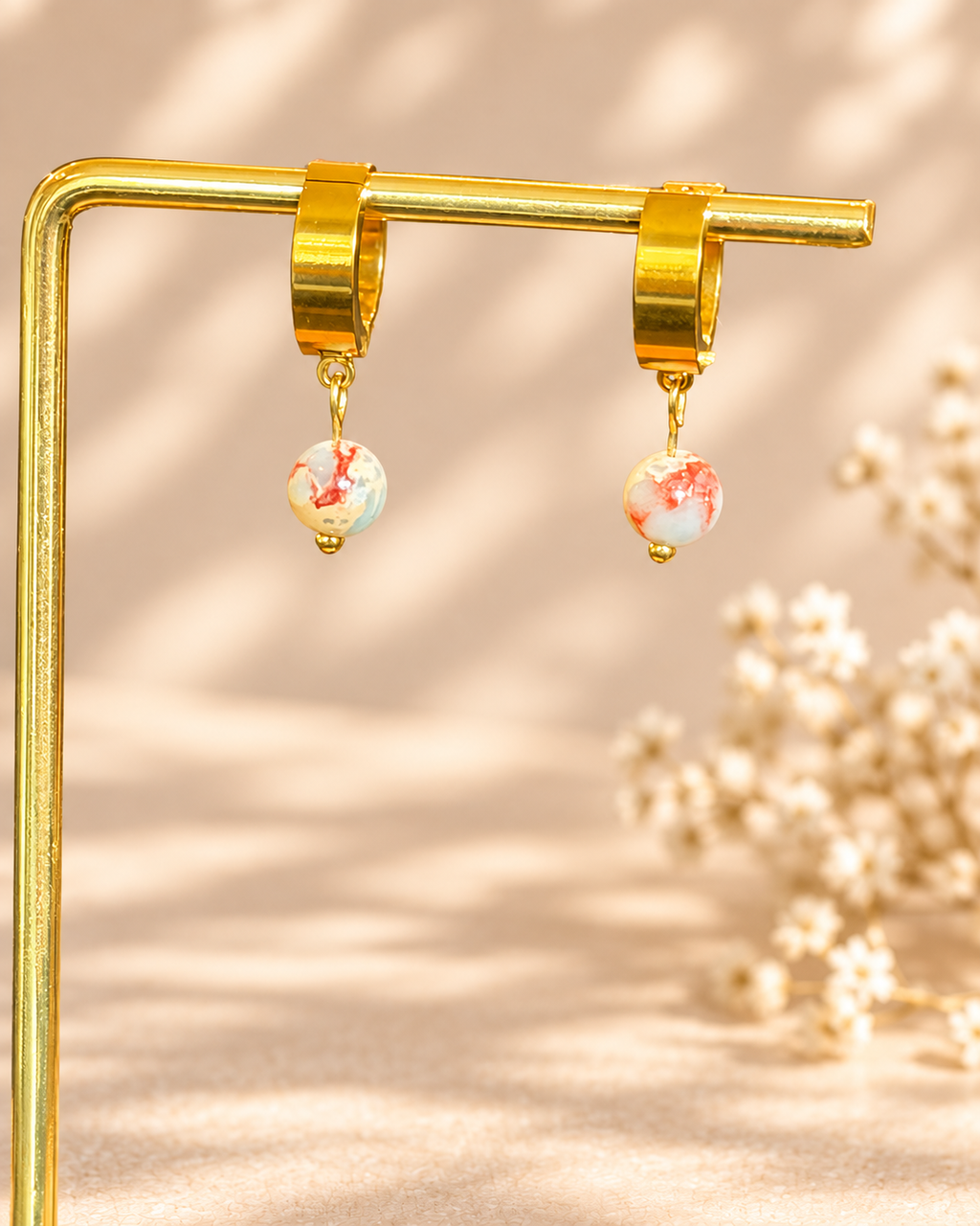 A pair of gold huggie hoop earrings with dangling marbled beads in white, red, and light blue, displayed on a gold stand against a warm beige background with soft floral bokeh.