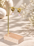 Gold hoop earrings with small iridescent pink and white bead drops, displayed on a gold stand with a wooden base against a warm beige background with soft floral props.