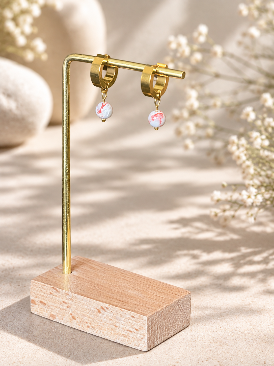 Gold hoop earrings with small iridescent pink and white bead drops, displayed on a gold stand with a wooden base against a warm beige background with soft floral props.