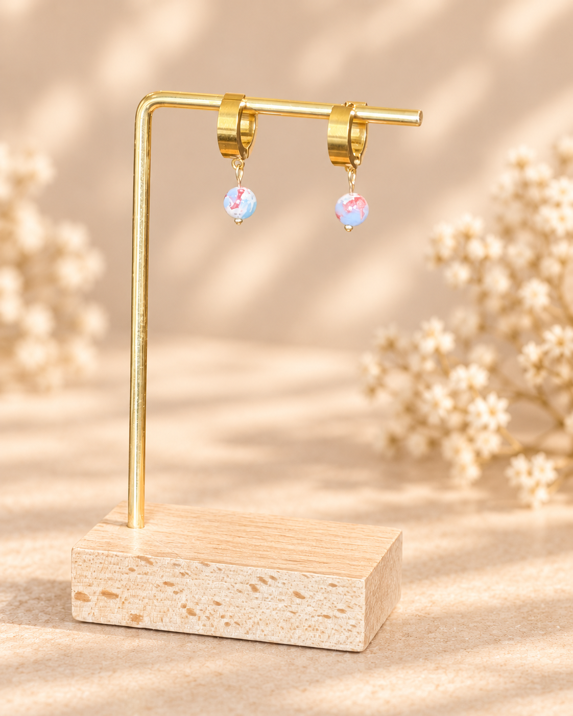 A pair of gold hoop earrings with small iridescent blue and pink teardrop charms, displayed on a gold stand with a wooden base against a warm beige background with soft floral bokeh.