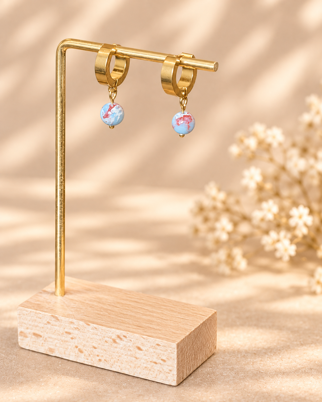 A pair of small hoop earrings with dangling round blue and pink marbled beads, displayed on a gold-tone stand with a wooden base against a warm beige background with soft floral decor.