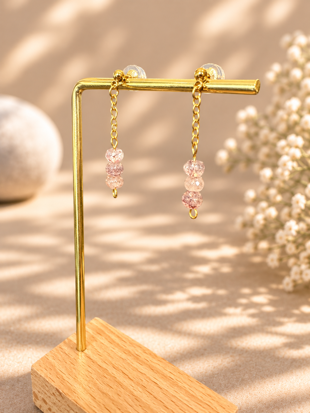 A pair of gold dangle earrings with pink faceted beads, displayed on a gold L-shaped stand over a wooden base against a warm beige background with soft shadows and dried flowers.