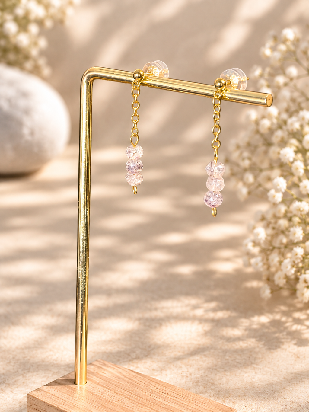 Gold-tone dangle earrings with clear faceted beads hanging from a gold display stand, set against a warm beige background with soft floral bokeh.