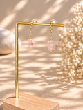Gold-tone dangle earrings with pink faceted beads, displayed on a gold stand over a wooden base against a warm beige background with soft shadows and dried flowers.
