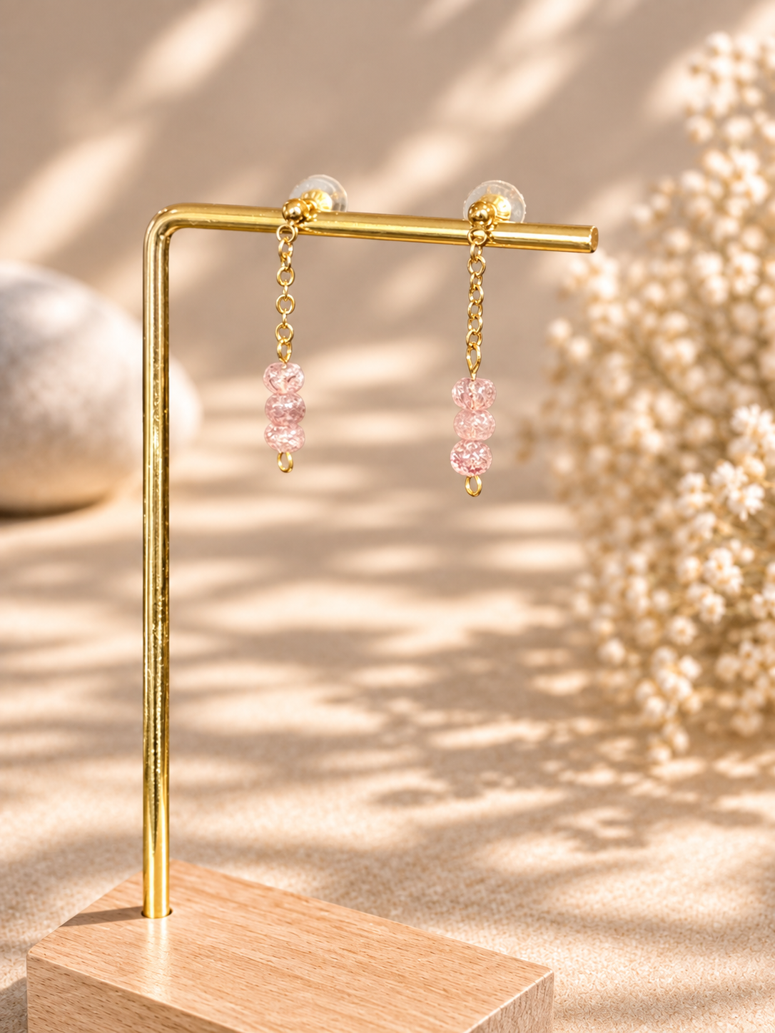 Gold-tone dangle earrings with pink faceted beads, displayed on a gold stand over a wooden base against a warm beige background with soft shadows and dried flowers.