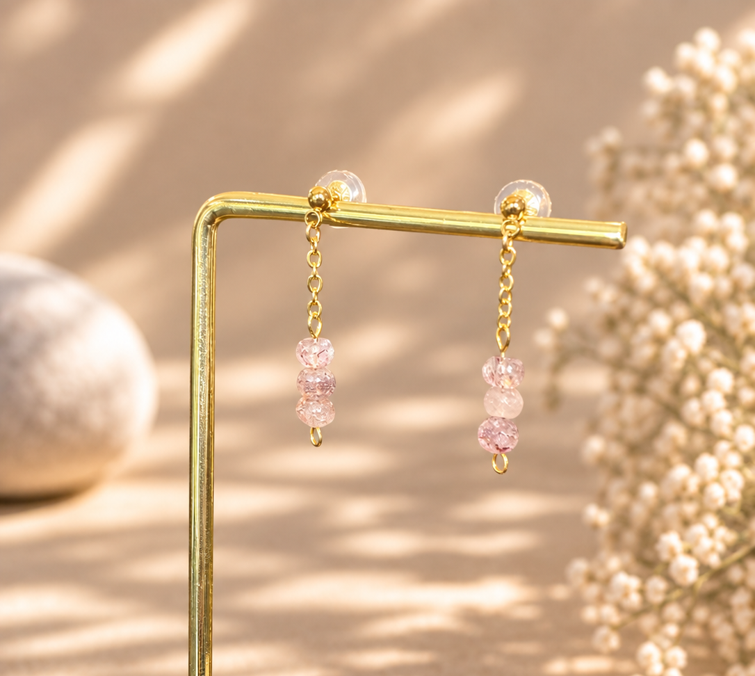 A pair of gold-tone dangle earrings with pink faceted beads, displayed on a gold L-shaped stand with a wooden base against a warm beige background with soft shadows and dried flowers.
