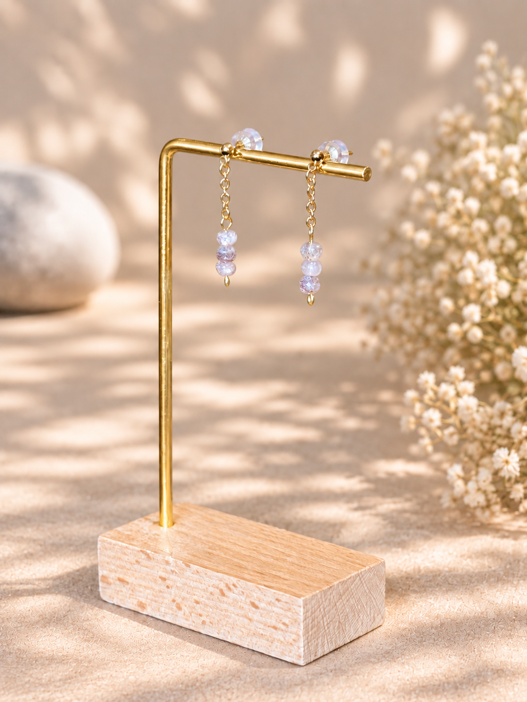 Gold-tone dangle earrings with clear faceted beads displayed on a small wooden stand with a wood base, set against a warm beige background with soft floral decor.