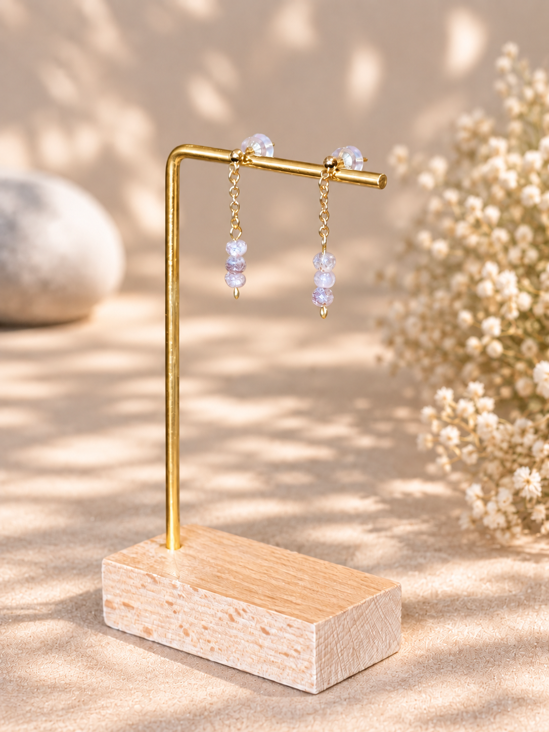 Gold-tone dangle earrings with clear faceted beads displayed on a small wooden stand with a wood base, set against a warm beige background with soft floral decor.