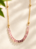 A delicate gold chain necklace featuring a curved row of faceted pink gemstone beads, displayed on a soft cream fabric background with soft leaf shadows.