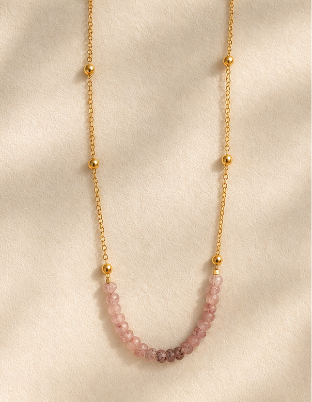 A delicate gold chain necklace with small gold beads and a curved section of pink faceted gemstones, displayed on a cream-colored jewelry stand with soft leaf shadows.