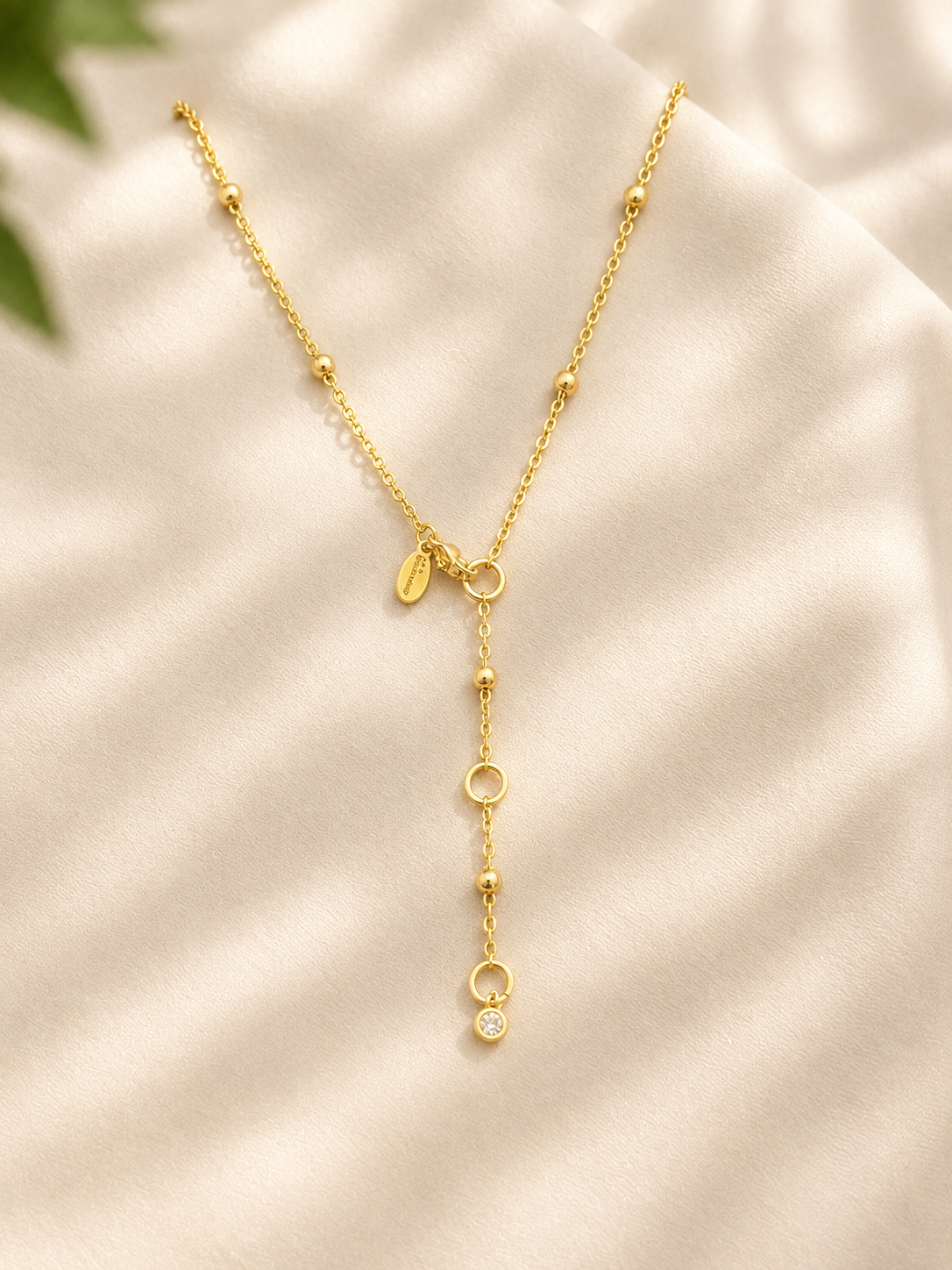 A delicate gold Y-necklace with small bead accents, open circle details, and a small clear stone pendant, displayed on a soft cream satin fabric background.