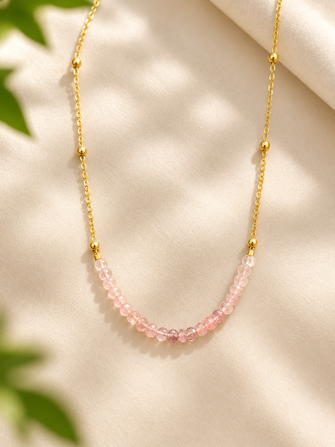 A delicate gold chain necklace featuring a curved section of small, faceted pink beads, displayed on a soft beige fabric background with subtle green leaf shadows.