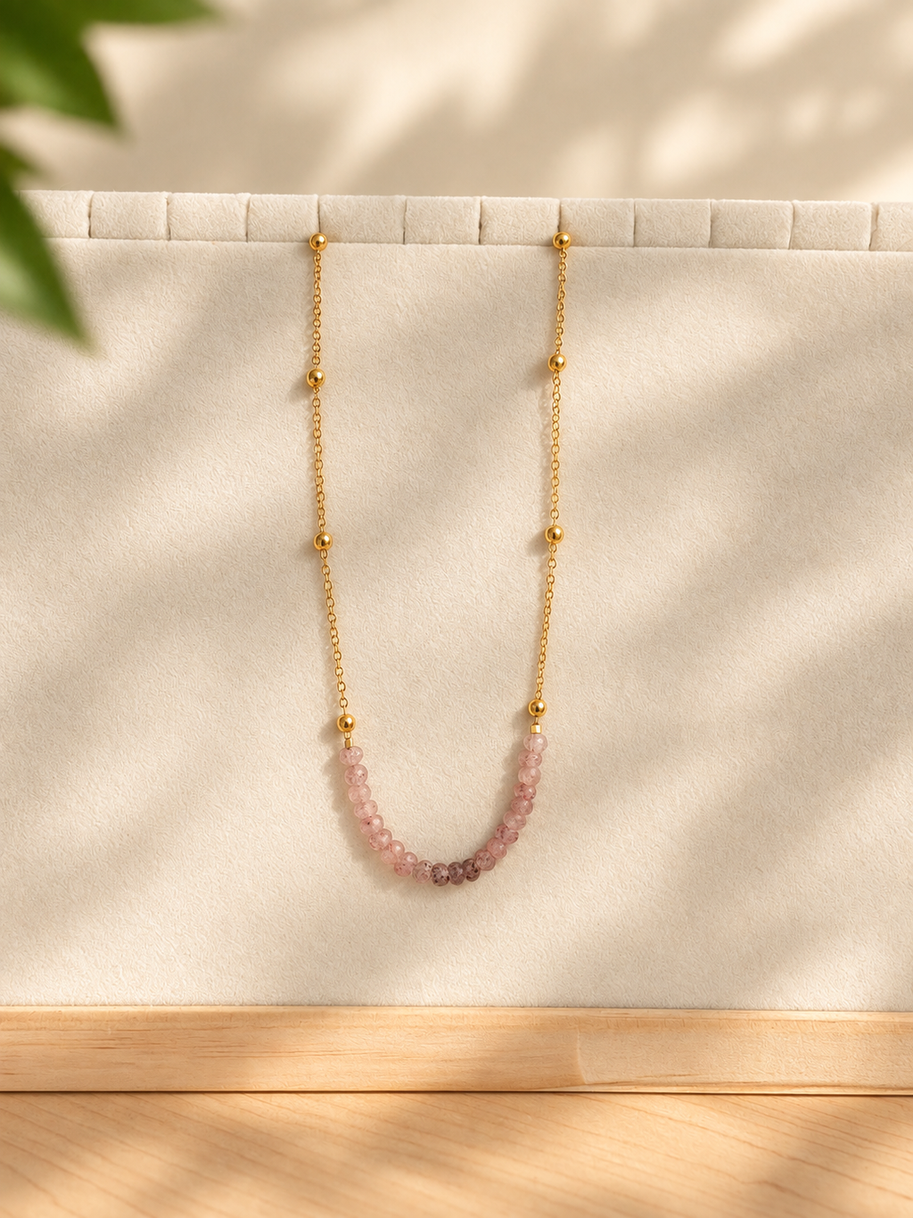 A delicate gold chain necklace with small gold beads and a curved section of pink faceted gemstones, displayed on a cream-colored jewelry stand with soft leaf shadows.
