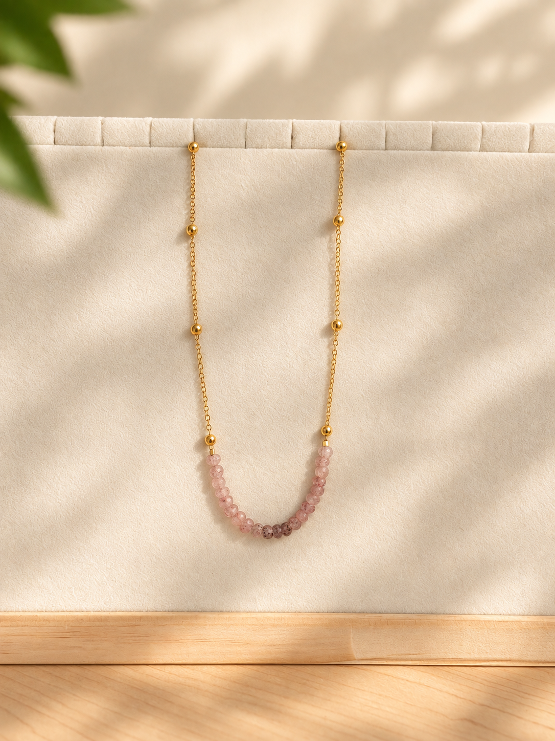 A delicate gold chain necklace with small gold beads and a curved section of pink faceted gemstones, displayed on a cream-colored jewelry stand with soft leaf shadows.