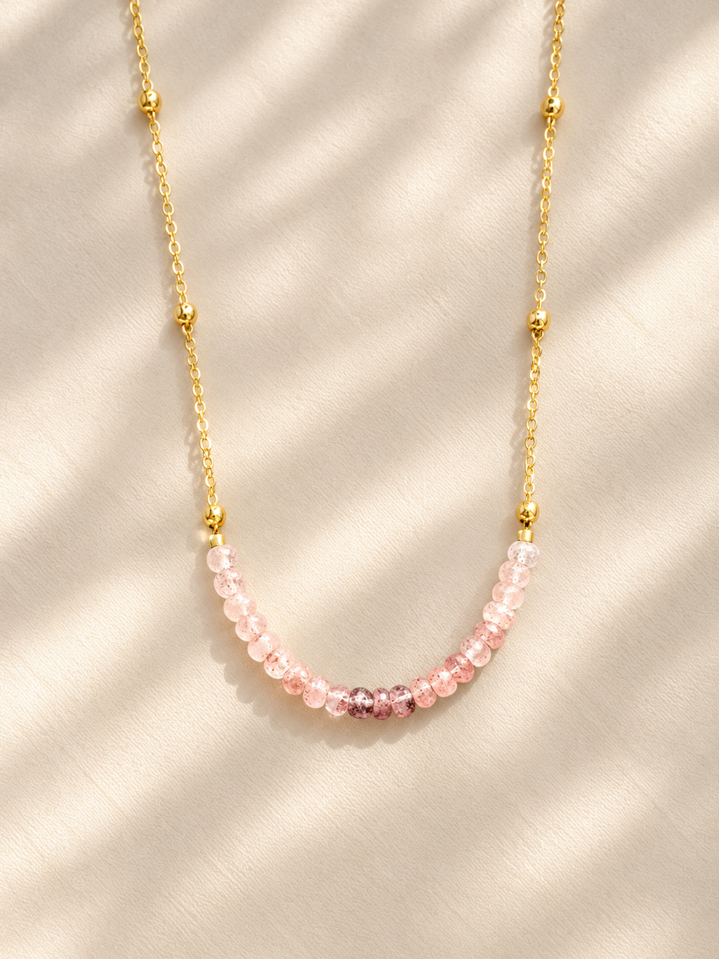 A delicate gold chain necklace featuring a curved row of faceted light pink beads, displayed on a textured cream fabric with soft shadow overlays.