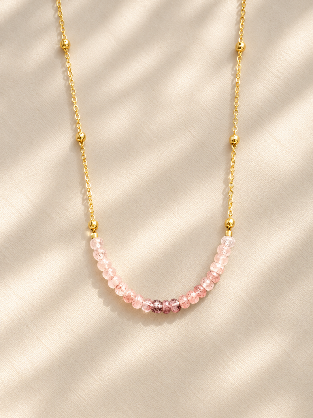 A delicate gold chain necklace featuring a curved row of faceted light pink beads, displayed on a textured cream fabric with soft shadow overlays.