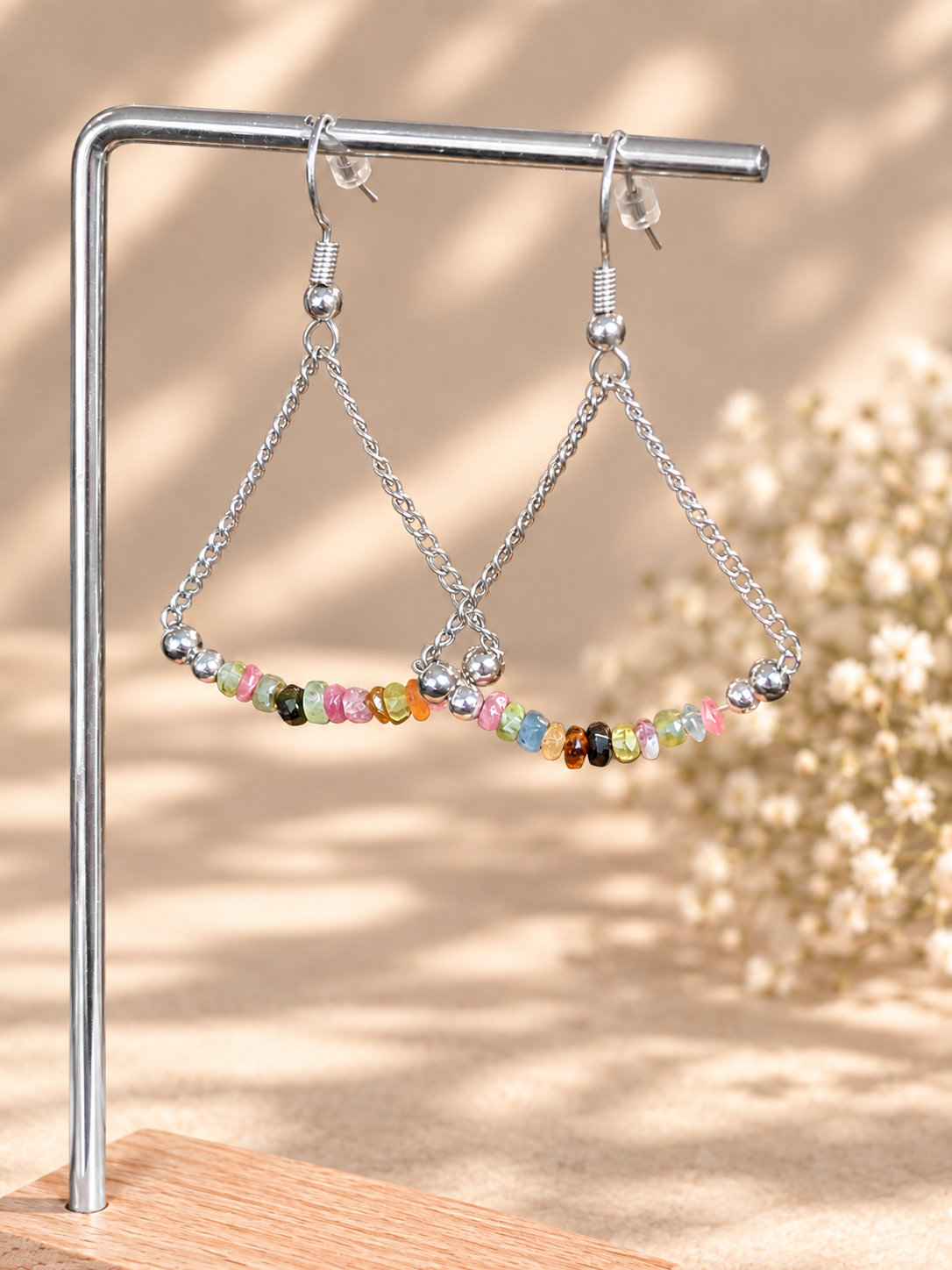Silver-tone triangle dangle earrings with small multicolored faceted beads, hanging from a metal display stand against a warm beige background with soft floral shadows.