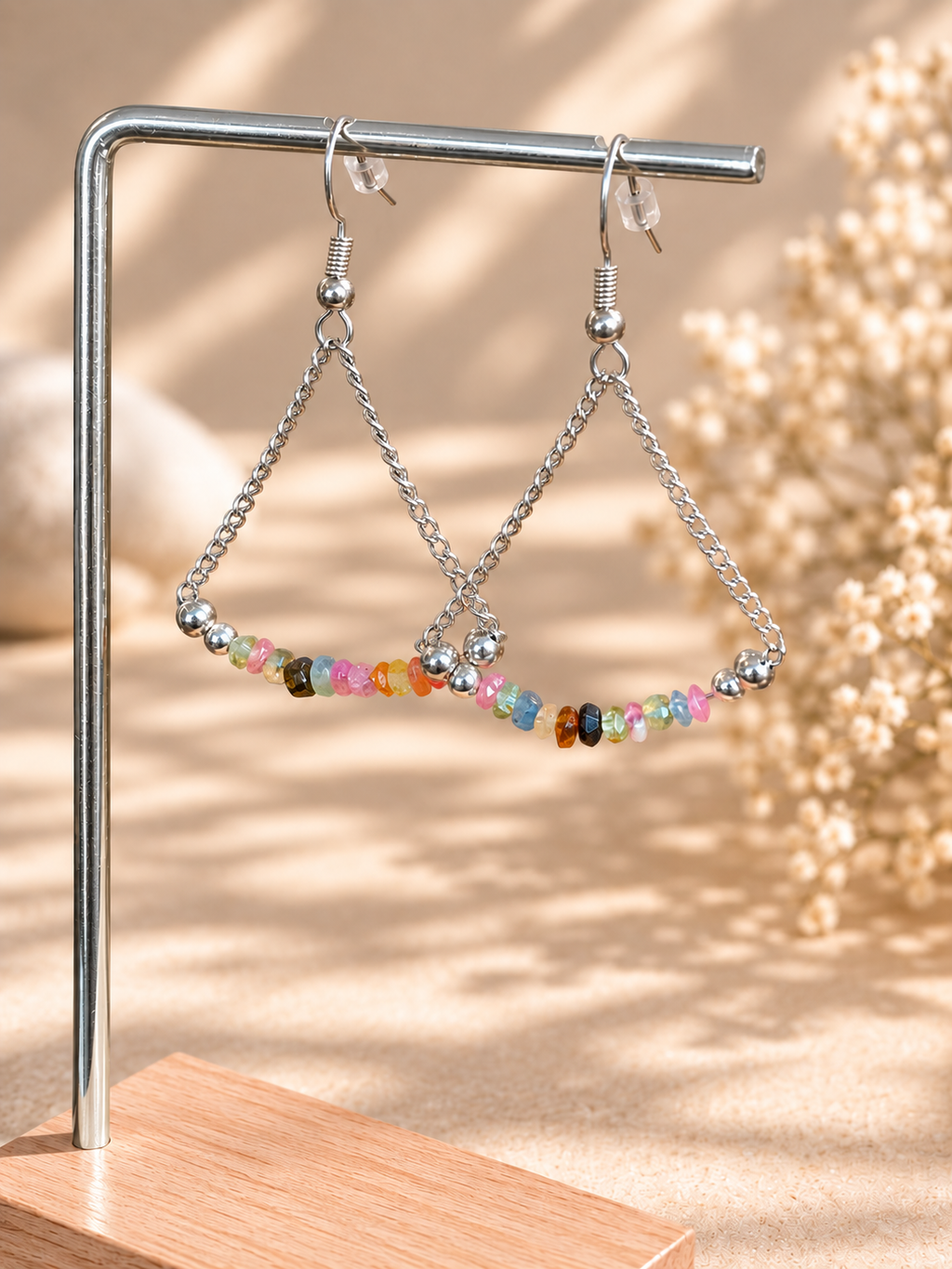 Silver-tone triangle dangle earrings with a row of small multicolored beads, displayed on a metal stand against a warm beige background with soft shadows.