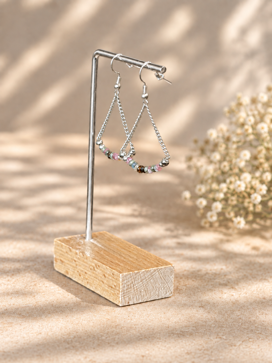 Silver-tone beaded hoop and chain earrings displayed on a small metal stand with a wooden base, set against a warm beige textured background with soft floral decor and dappled sunlight.