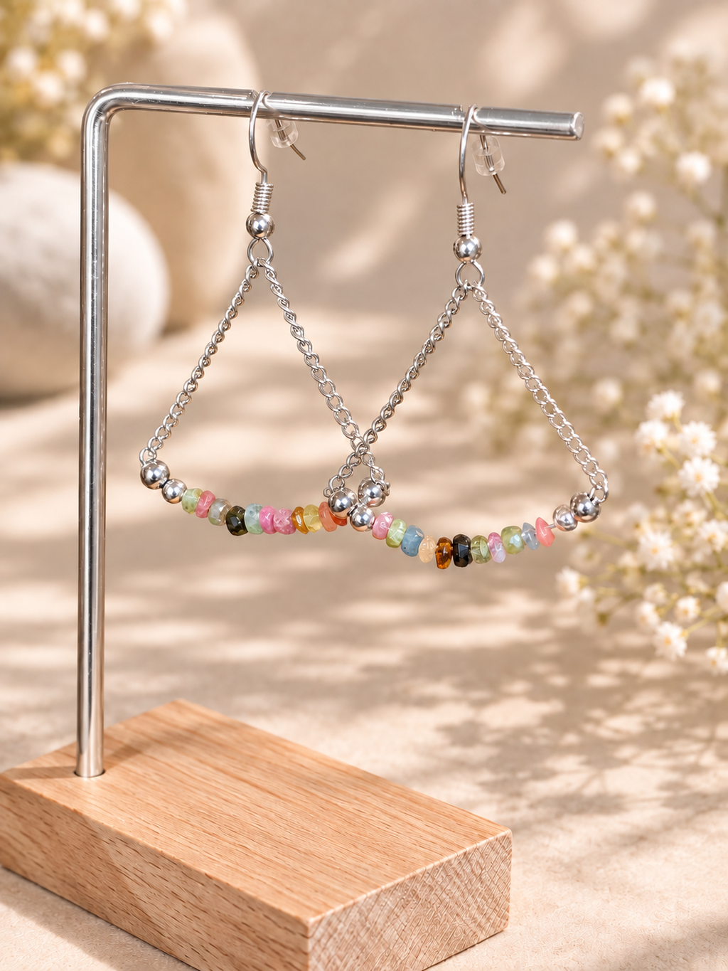 Silver-tone triangle dangle earrings with small multicolored beads, displayed on a metal stand with a wooden base against a softly blurred beige background with dried flowers.
