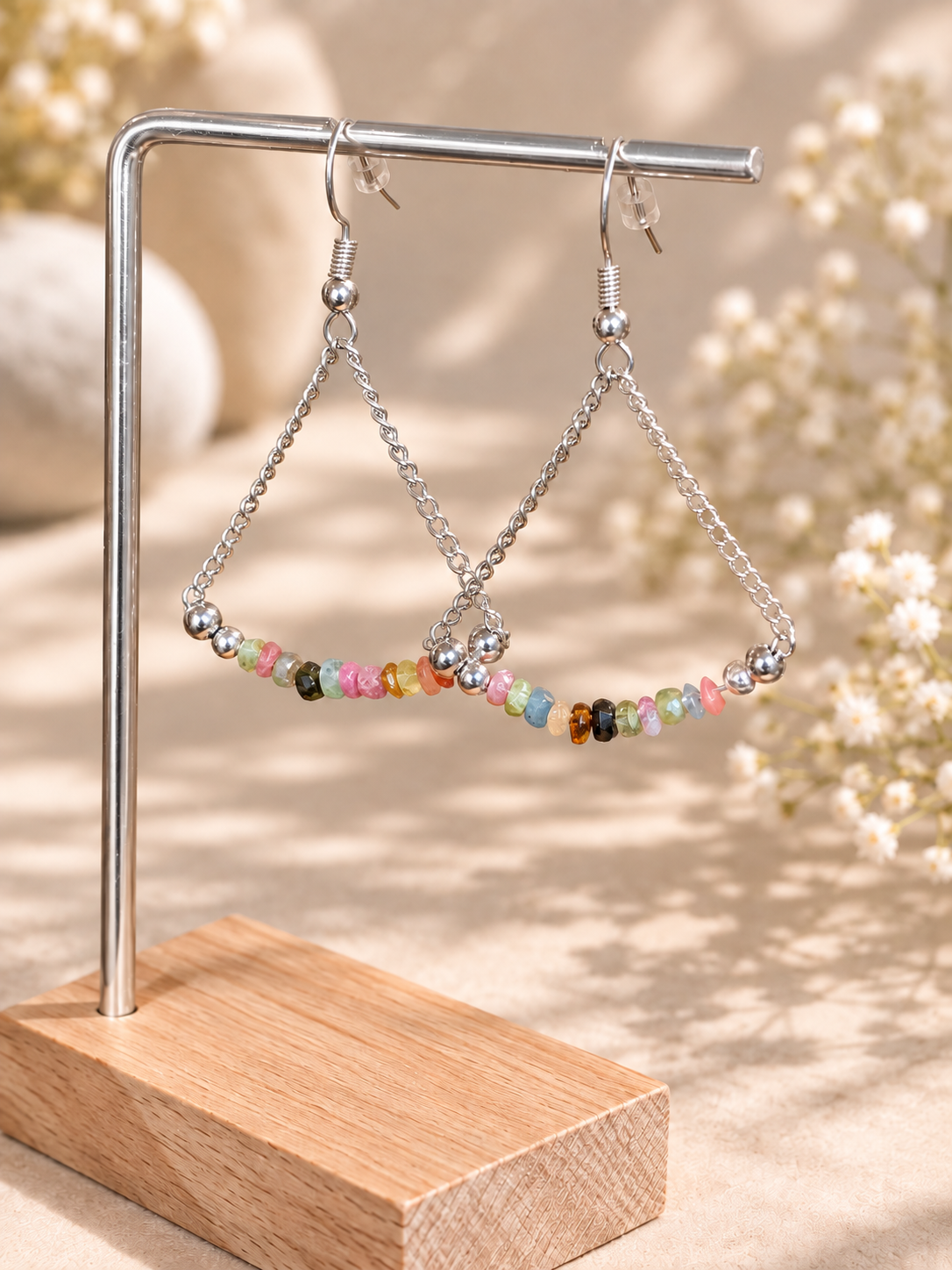 Silver-tone triangle dangle earrings with small multicolored beads, displayed on a metal stand with a wooden base against a softly blurred beige background with dried flowers.