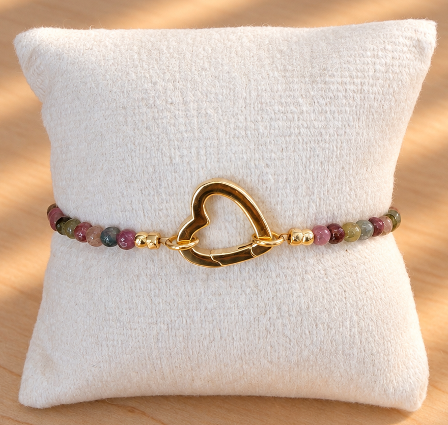 A beaded bracelet with a polished gold heart-shaped clasp, displayed on a white fabric jewelry pillow over a light wood surface with warm natural shadows.