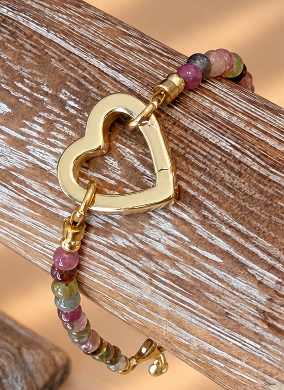 A gold heart-shaped clasp bracelet with multi-colored gemstone beads, displayed on a rustic wooden block.