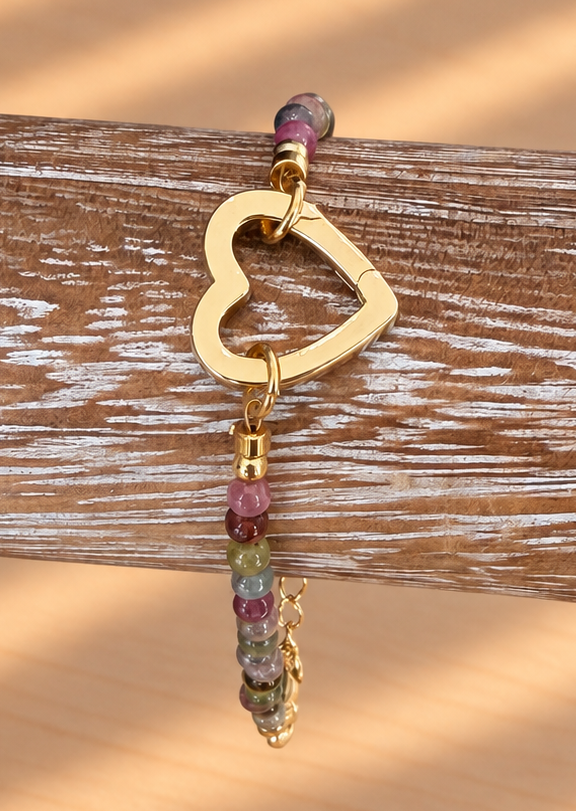 A gold heart-shaped clasp with a dangling beaded charm in purple, green, and gold, displayed on a rustic wooden stand against a warm wooden background with soft diagonal shadows.