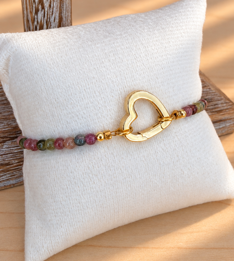 A beaded bracelet with a gold heart-shaped clasp resting on a white fabric jewelry pillow over a light wood surface.