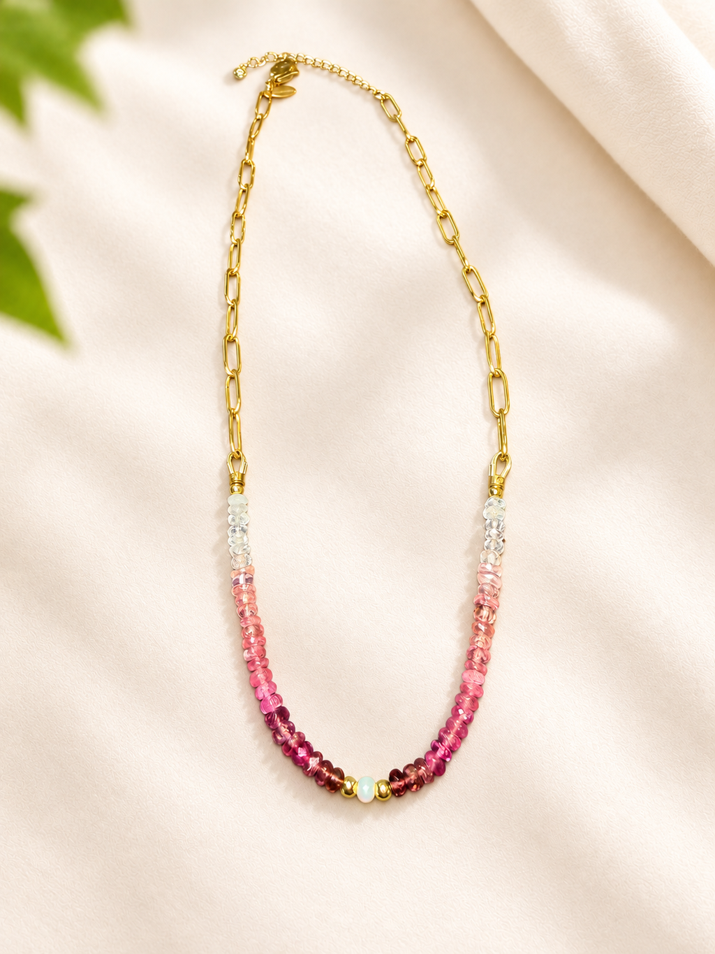 A gold chain necklace with a beaded drop section transitioning from clear faceted beads at the top to vibrant pink and magenta beads at the bottom, displayed on a soft cream fabric background.