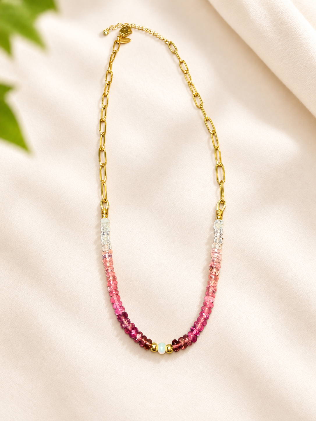 A gold chain necklace with a beaded drop section transitioning from clear faceted beads at the top to vibrant pink and magenta beads at the bottom, displayed on a soft cream fabric background.